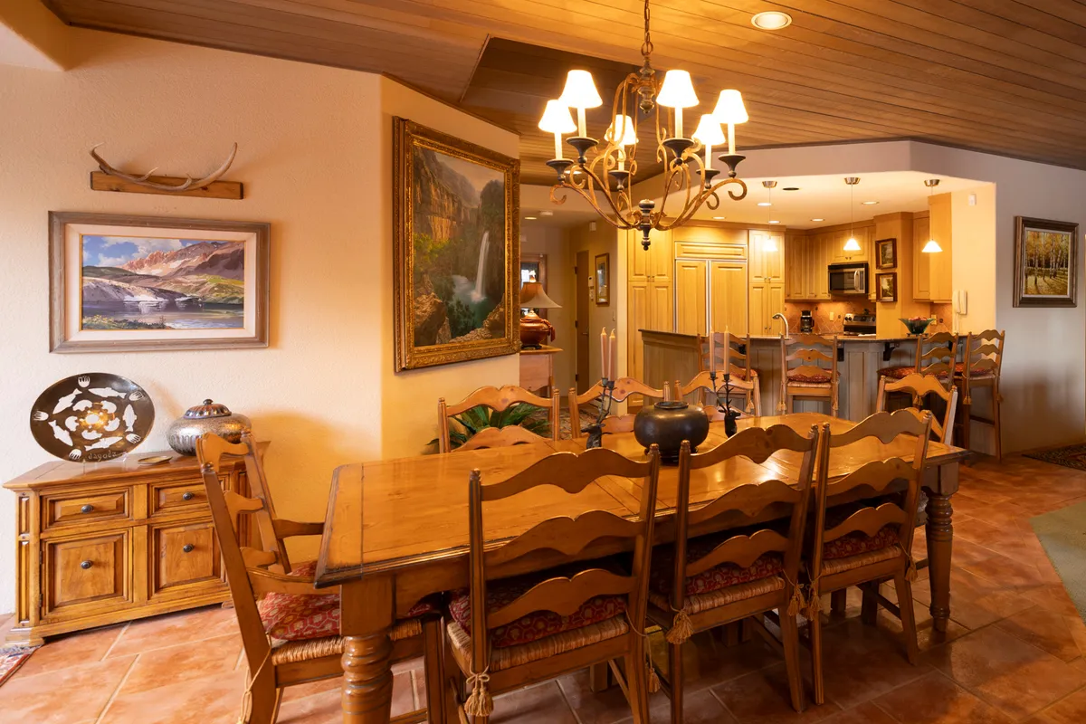 The image showcases a spacious, open-concept dining area with a large wooden dining table set beneath a chandelier. Paintings adorn the walls, and the kitchen area is visible in the background, emphasizing the open layout. The space is designed for gatherings, celebrations, and shared meals.