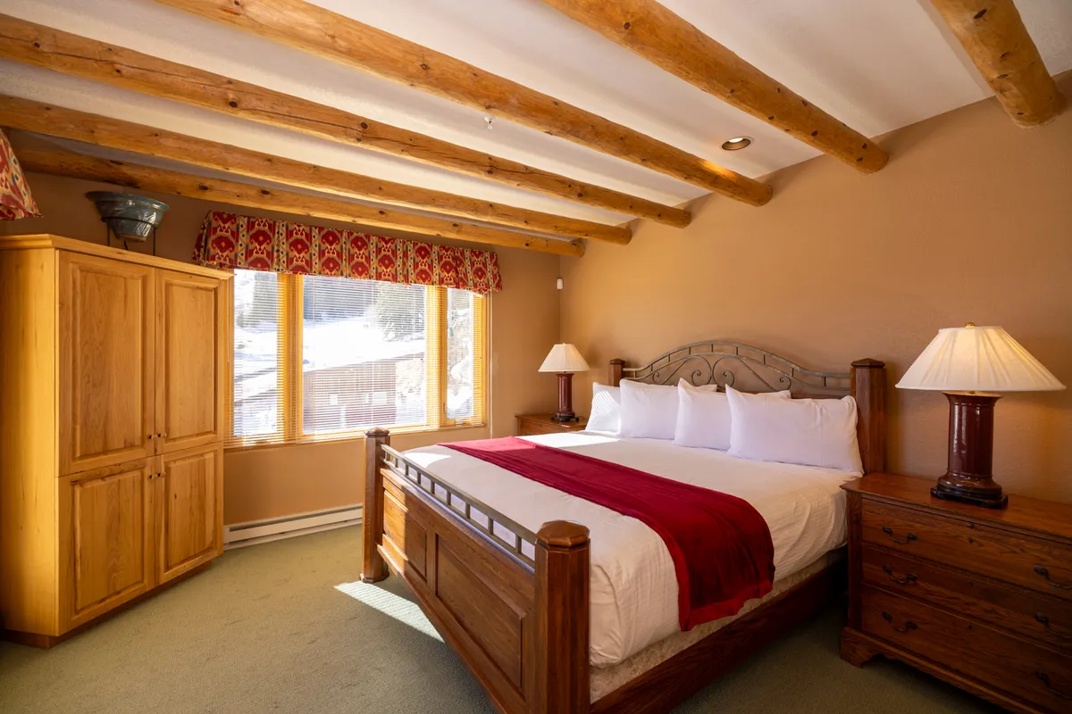 A spacious bedroom with a large wooden king bed, flooded with natural light from a window. Additional details include a wooden wardrobe and a stylish decorative lamp, creating a tranquil, inviting atmosphere ideal for relaxation.