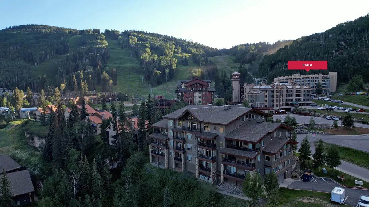 A sweeping exterior shot of Purgatory Village, showcasing residential buildings with a backdrop of majestic mountains and a visible parking area. The scene emphasizes accessibility and the ease of moving between social resort activities and private comfort.
