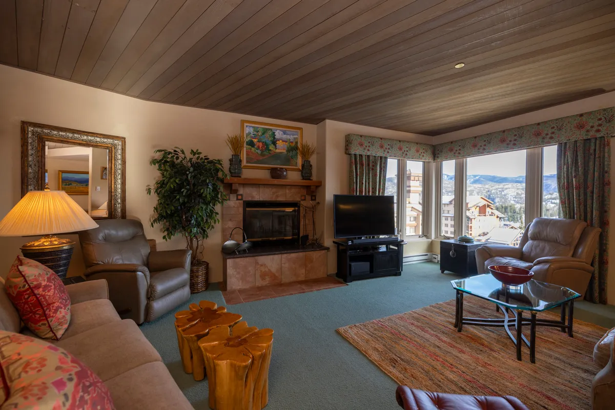 A cozy open-plan living area featuring a glowing fireplace surrounded by plush seating, large windows that frame breathtaking mountain views, and wooden furniture. The inviting space is arranged for socializing and relaxing, with the warmth of the fire and the natural light creating a welcoming retreat after a day on the slopes.