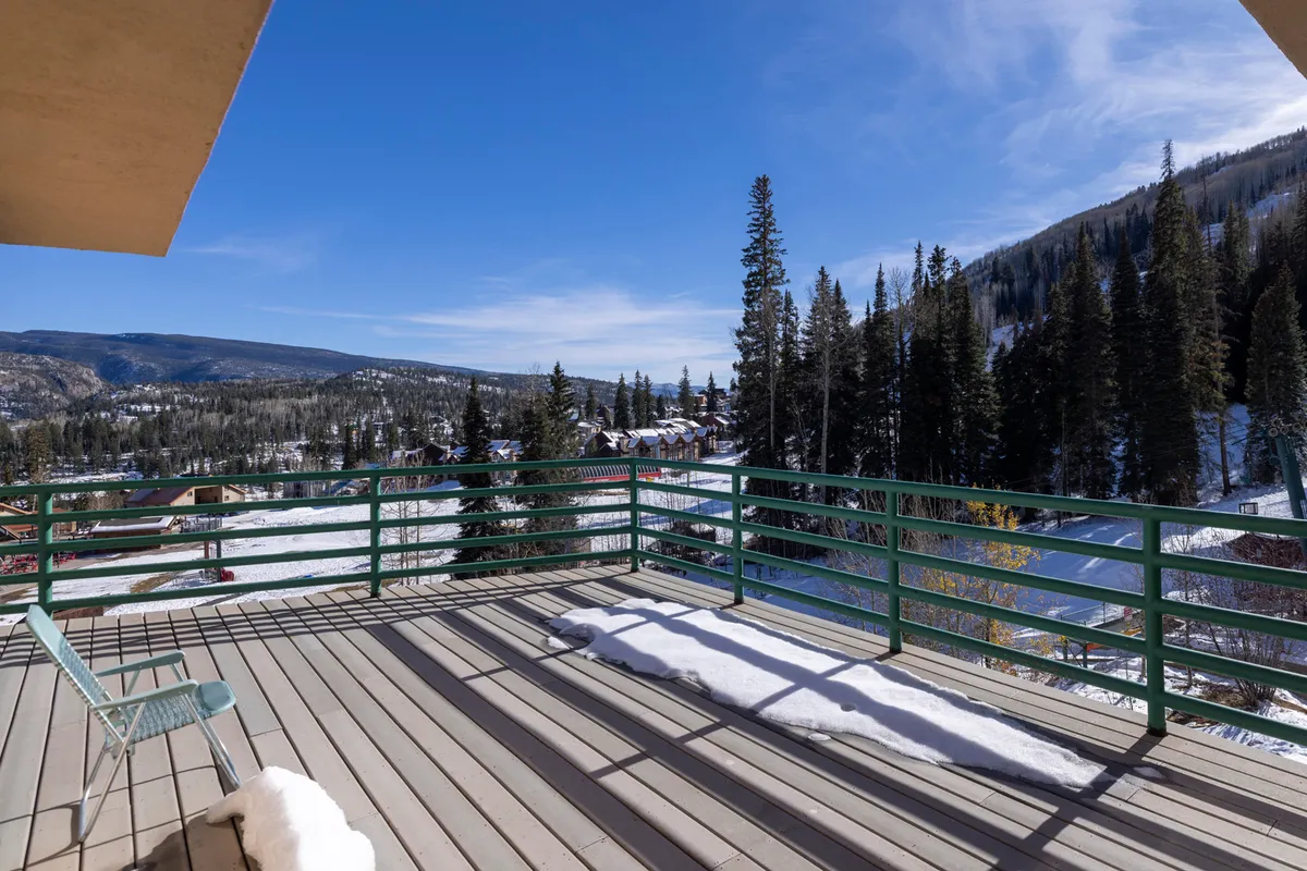 The image showcases a snow-covered landscape with tall pine trees and a breathtaking panoramic view of the sun rising over the Needles Mountains. The balcony railing suggests a vantage point perfect for soaking in the scenery. Everything is bathed in peaceful morning light, evoking tranquility and the promise of adventure.