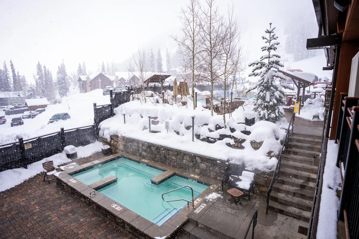 An inviting exterior view of a steaming hot tub surrounded by snow, pine trees, and a rustic stone wall. The backdrop features a peaceful winter landscape, suggesting relaxation and camaraderie after a day of outdoor activity.