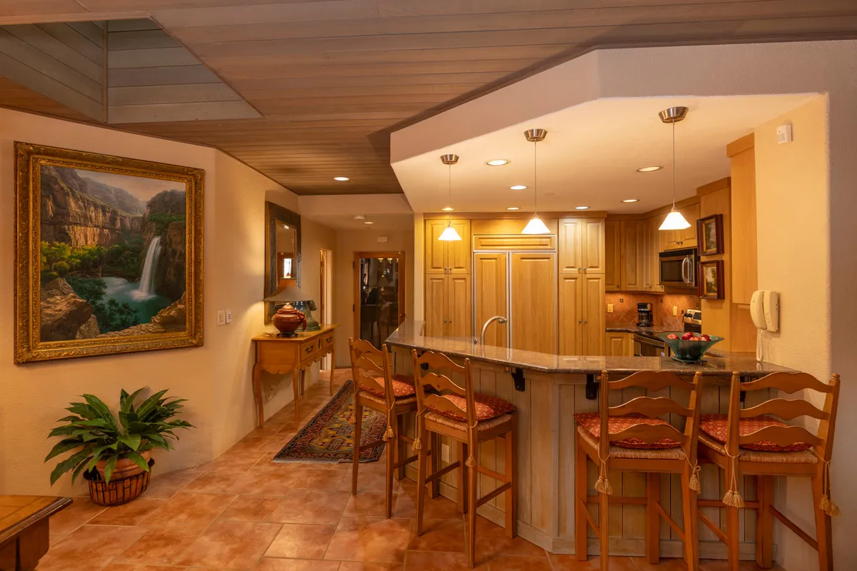 This interior shot features a modern kitchen with a central island, wooden cabinetry, and bar stools. A framed landscape painting adds a touch of warmth. The space is clearly designed for both preparation and socializing, emphasizing the kitchen as a gathering place.