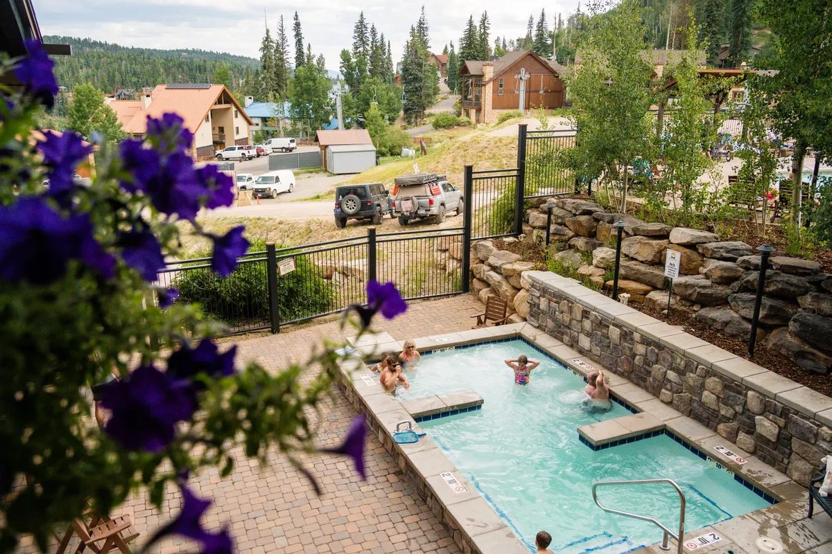 A cheerful outdoor scene showcasing a crystal-clear pool and adjacent hot tub, set against the natural splendor of surrounding mountains and colorful flowers. The area is designed for both relaxation and socializing, harmoniously blending resort amenities with the beauty of nature.