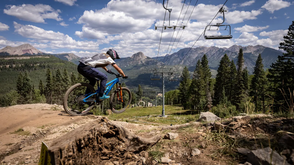 A dynamic scene showing a mountain biker navigating a rugged trail with a panoramic view of mountain peaks, ski lift cables, and dense pine forests. The sky is clear and blue, infusing the image with energy and highlighting the summer adventure possibilities right outside the resort.