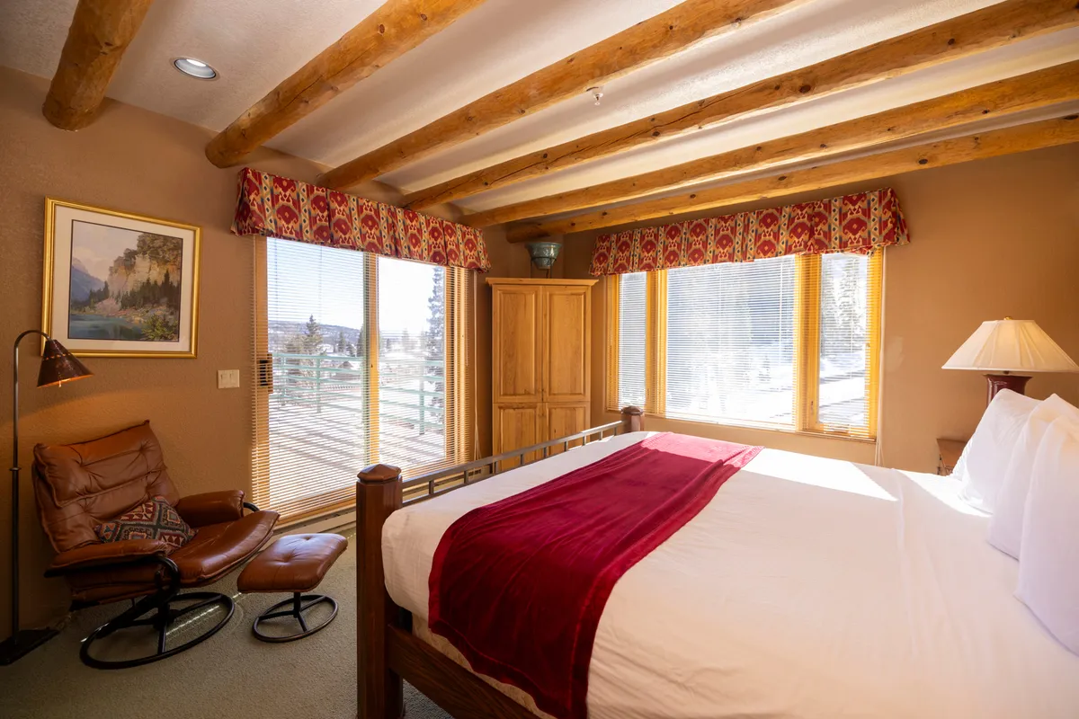 A serene bedroom with a bed dressed in a red accent blanket, large windows allowing in plenty of natural light, exposed wooden beams, and a comfortable chair positioned by the window. The space feels both tranquil and inviting—ideal for peaceful mornings or stargazing at night.