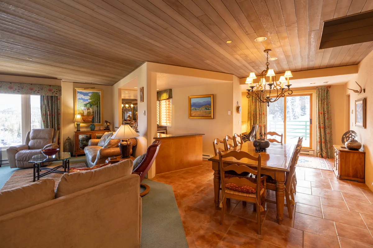 A cozy dining area featuring a wooden dining table, elegant chandelier, a nearby sofa for relaxed conversation, and large windows allowing natural light to pour in. The space invites lingering over meals and connects visually to the living area, suggesting an easy transition between spaces.