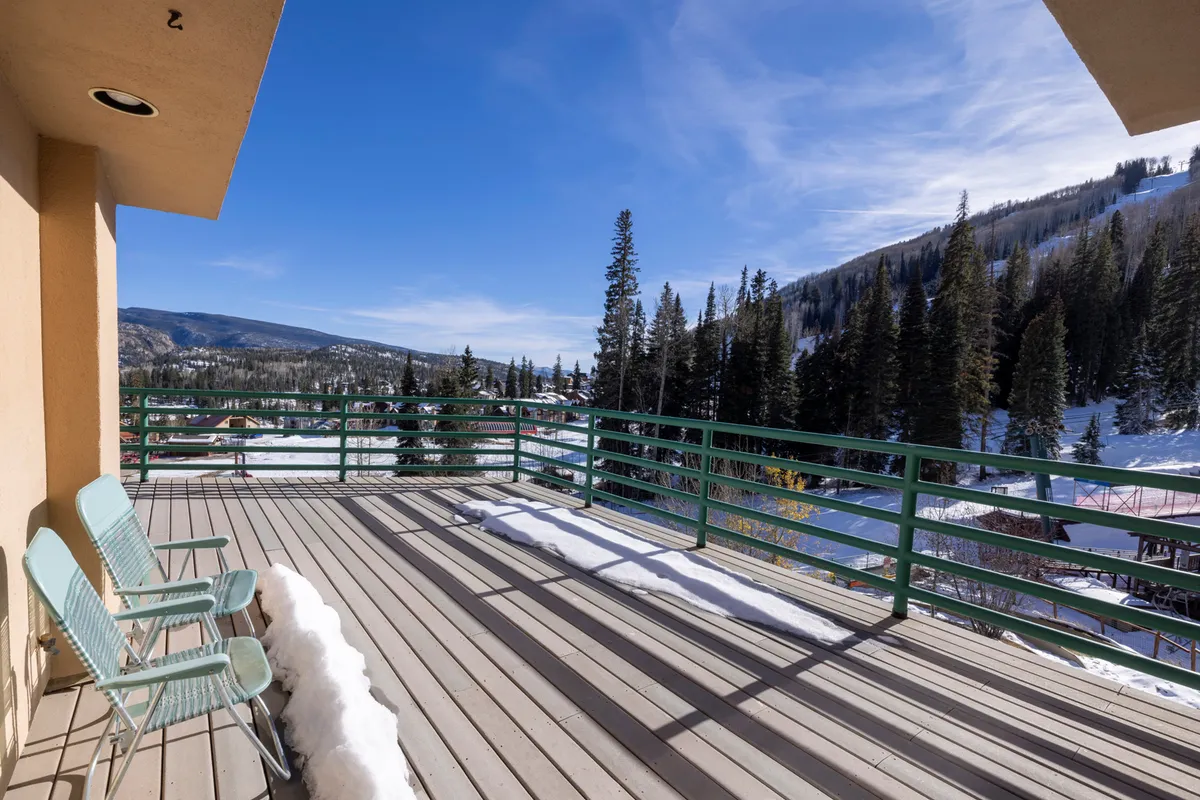 This image captures a serene private deck area featuring two green chairs, a metal railing, snow-covered ground, and breathtaking distant mountain views against a backdrop of pine trees. The scene evokes the tranquil experience of enjoying coffee or unwinding while surrounded by alpine scenery.