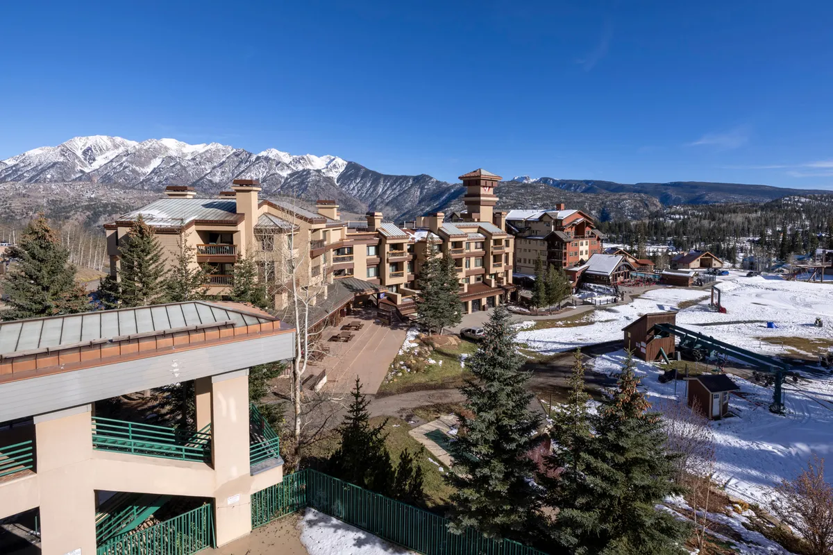 A stunning exterior shot of the ski resort nestled in a snow-covered landscape, set against a dramatic mountain range and dotted with evergreen pine trees. The scene captures both the proximity of the resort to the slopes and the breathtaking Needles Mountains beyond, evoking a sense of adventure and alpine serenity.