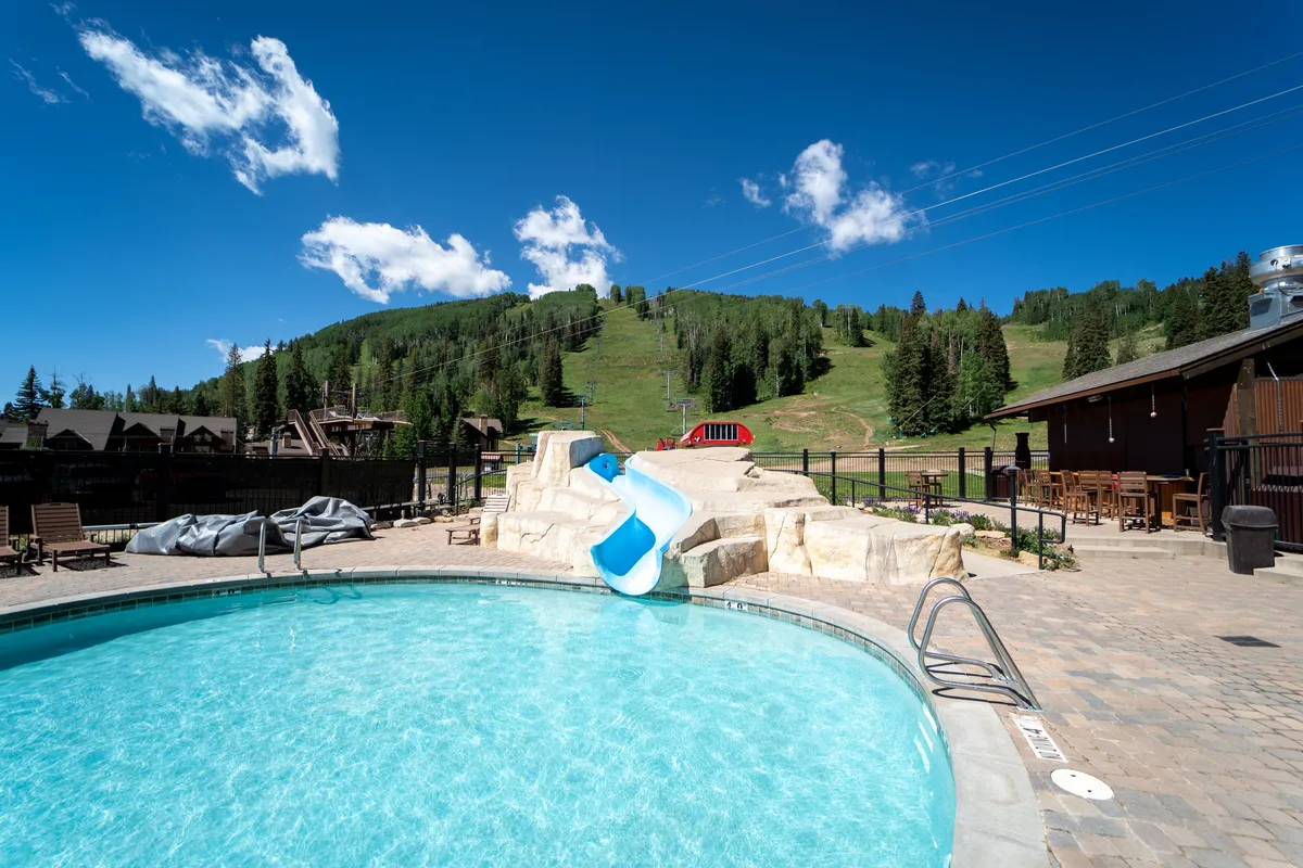 An outdoor swimming pool with a water slide, surrounded by mountains. The pool area is lively with families enjoying a sunny day, set against a spectacular mountain backdrop.